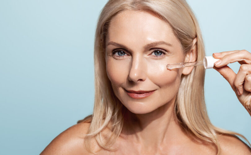 Porträt einer älteren Frau, die ihre Haut mit einem kosmetischen Gesichtswasser und einem Aha-Serum mit Hyaluronsäure zur Hautstraffung und für den Anti-Aging-Effekt pflegt, hält eine Pipette in der Nähe des Gesichts, blauer Hintergrund.