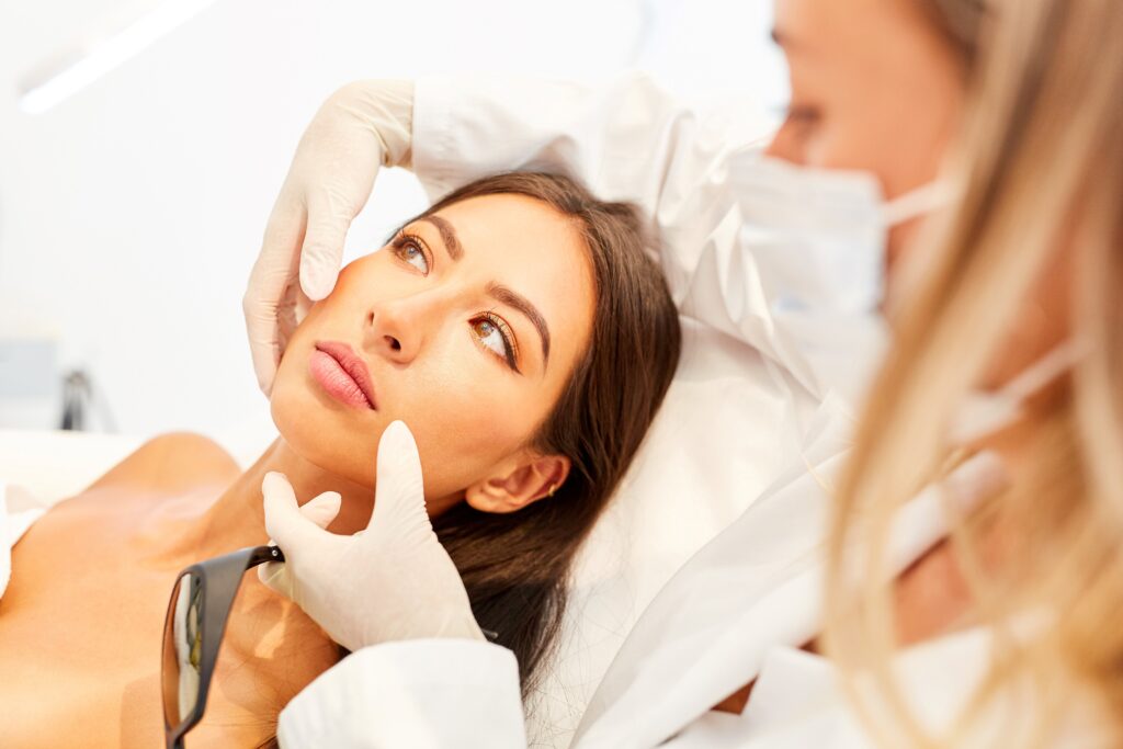 Kosmetikerin untersucht das Gesicht einer Patientin während der Vorbereitung zur Laser-Haarentfernung, beide tragen Masken.