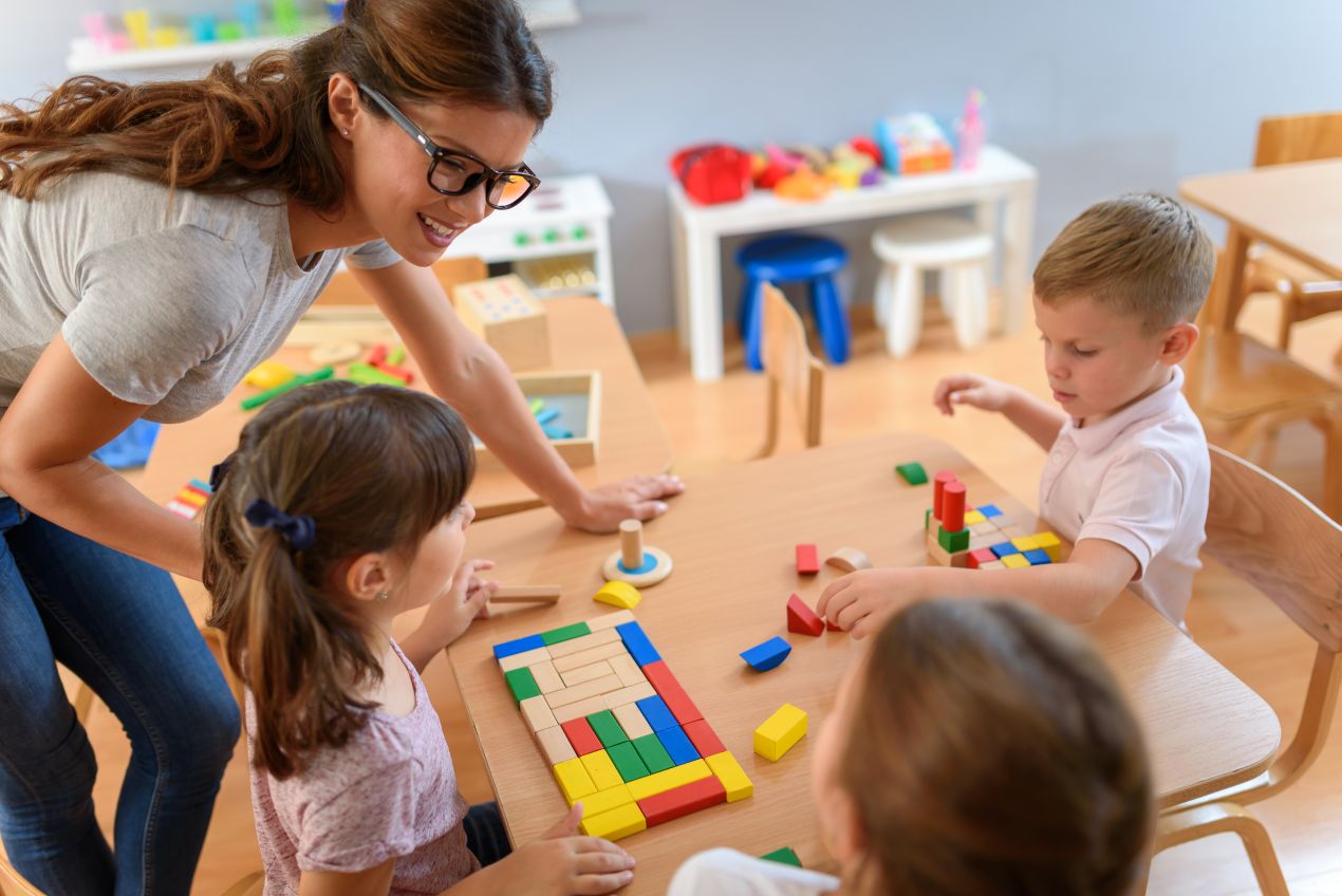 Eine Frau betreut spielende Kinder an einem Tisch mit Bauklötzen – Einblick in den Alltag fuer Quereinsteiger Kindergarten