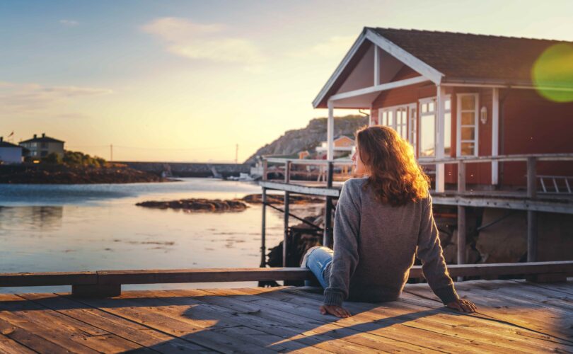 Schöne junge Reisende sitzt auf einer Holzpier vor dem Hintergrund eines traditionellen roten Rorbues auf dem Weg zu den Lofoten in Norwegen.