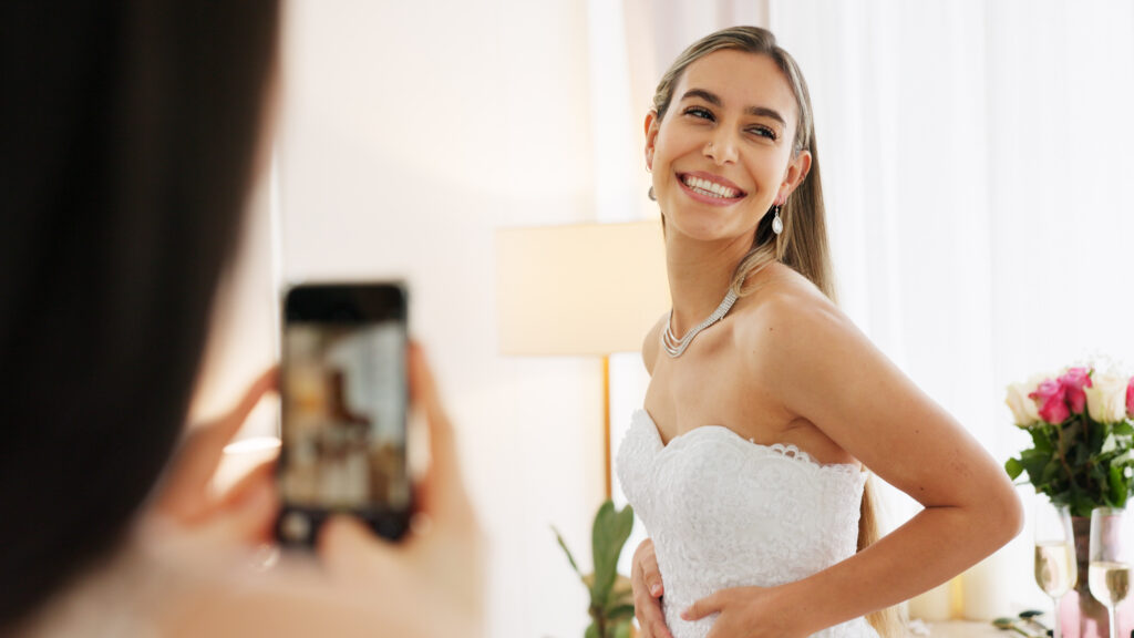 Lächelnde Frau in einem weißen Hochzeitskleid bei der Anprobe in Hamburg, eine Freundin fotografiert sie mit dem Smartphone