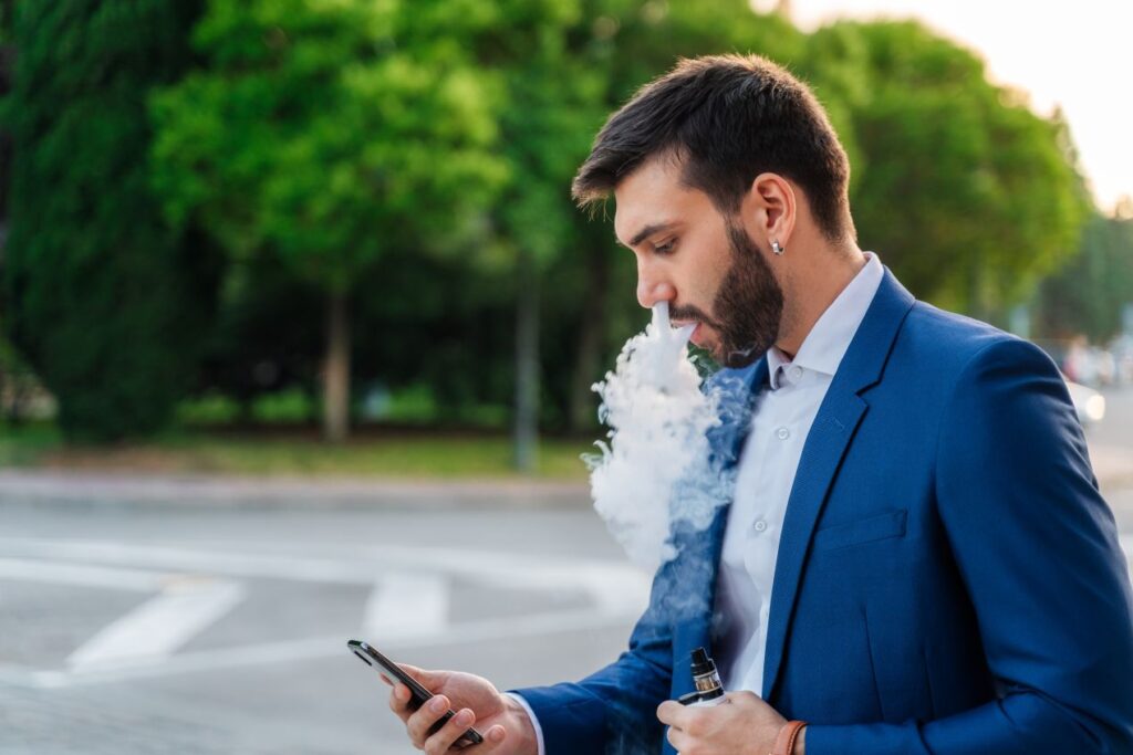 Mann im blauen Anzug dampft Elfbar Liquids auf der Strasse und schaut auf sein Smartphone