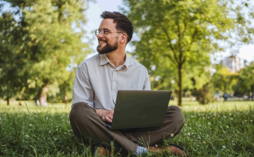 Geschäftsmann sitzt auf dem Gras in der Natur und nutzt einen Laptop zur Arbeit oder zur Freizeit.